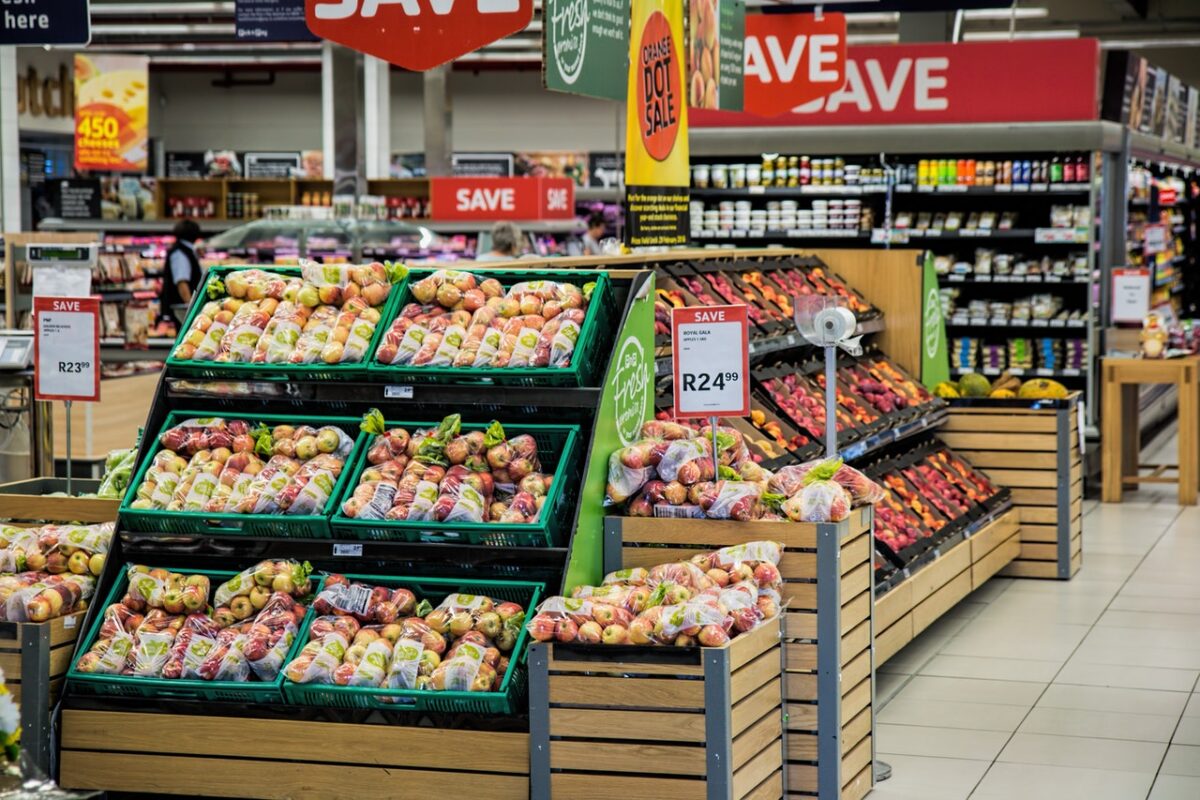 A brightly lit supermarket aisle featuring neatly organized shelves full of fresh fruits in baskets, with promotional signs offering discounts visible above and cautionary notices for potential supermarket accidents.