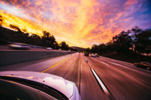 A dynamic, motion-blurred view of a highway from a car's hood, featuring other vehicles, construction accidents and a vivid sunset with dramatic orange and pink skies.