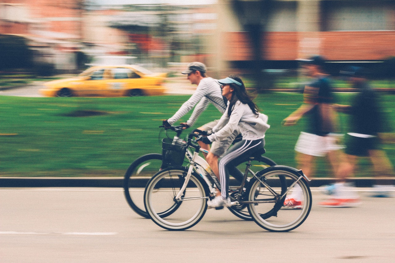 Two people cycling in an urban park, complying with bicycle helmet laws, with a blurred background showing motion, alongside someone jogging and a yellow taxi in the distance.