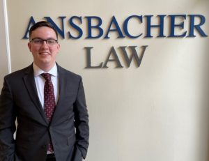 A professional young man in a suit and tie, one of the summer law clerks, smiling in front of a sign that reads "Ansbacher Law.