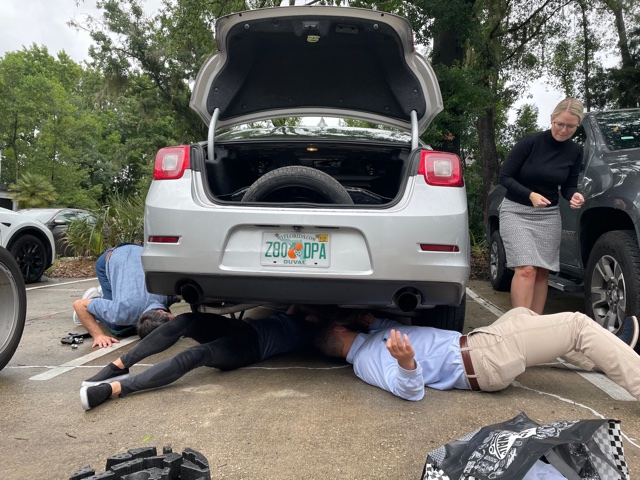 Three people lie on the ground under the rear of a silver car, inspecting it for Ansbacher Law, while a woman stands nearby, all dressed formally. The car’s trunk is open.