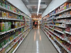 Aisle in a grocery store stocked with a variety of beverages on both sides, featuring a person with a shopping cart at the far end under bright fluorescent lights.