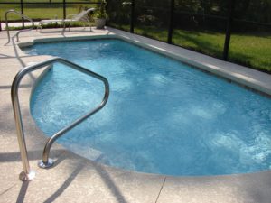 A backyard swimming pool with crystal clear blue water, prone to swimming pool accidents, surrounded by a concrete deck and metal fence, featuring shiny stainless steel handrails by the steps.