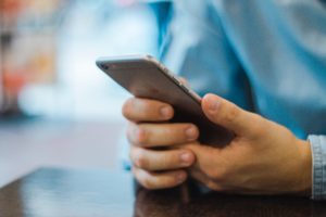 Close-up of a person's hands holding a smartphone at a table, scrolling through social media, with a blurred background focusing on the device and hands.