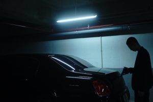 A silhouette of a man in a dimly lit garage, standing next to a parked car, seemingly opening its fuel or charging port under the inadequate glow of overhead lights.