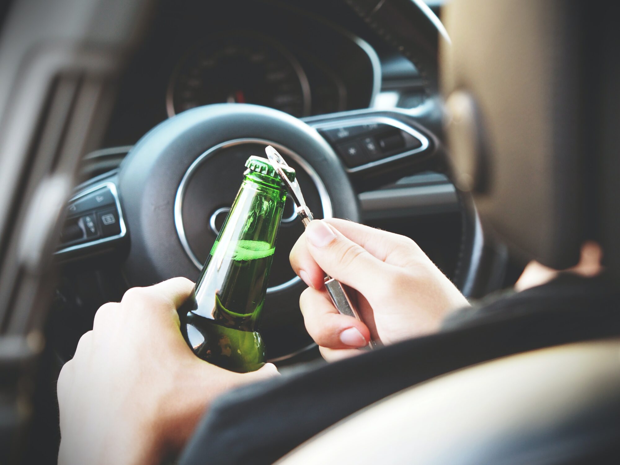 Driver holding a green bottle with a metallic opener attached to it, positioned near a car steering wheel, demonstrating drunk driving behavior.