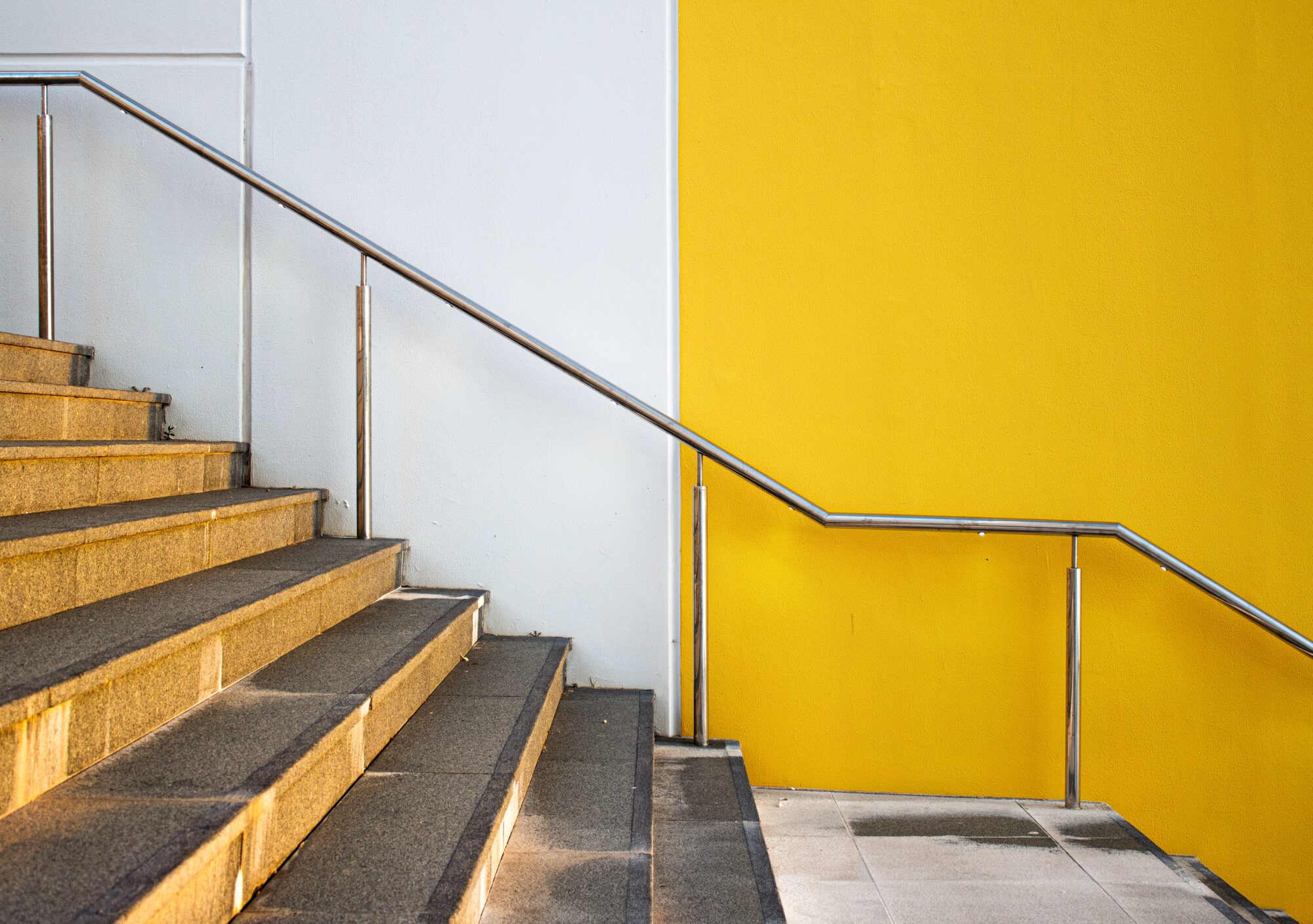 A stairway with textured golden steps and a metal handrail, beside a vibrant yellow wall and prone to stairwell accidents, contrasting with a white tiled floor, creating a bold architectural contrast.