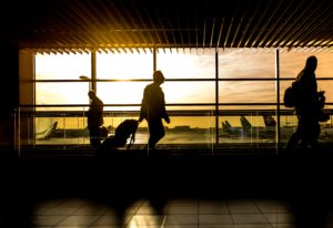 Silhouetted figures walking past caution signs inside an airport terminal at sunset, with airplanes visible through the large windows illuminated by golden sunlight.