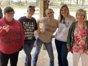 Five women pose smiling together outdoors, three younger and two older, wearing casual attire with some in festive-themed shirts for the 2021 Ugly Sweater Contest.