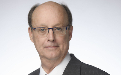 A middle-aged man with glasses, wearing a dark suit and tie, posing against a light gray background as part of a comprehensive plan. He has a serious expression and is looking directly at the camera.