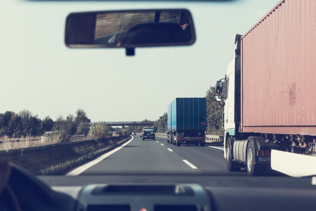 View from a car's interior showing a highway with large trucks in adjacent lanes under a clear sky, seen through the windshield with a rearview mirror at the top, highlighting areas prone to truck