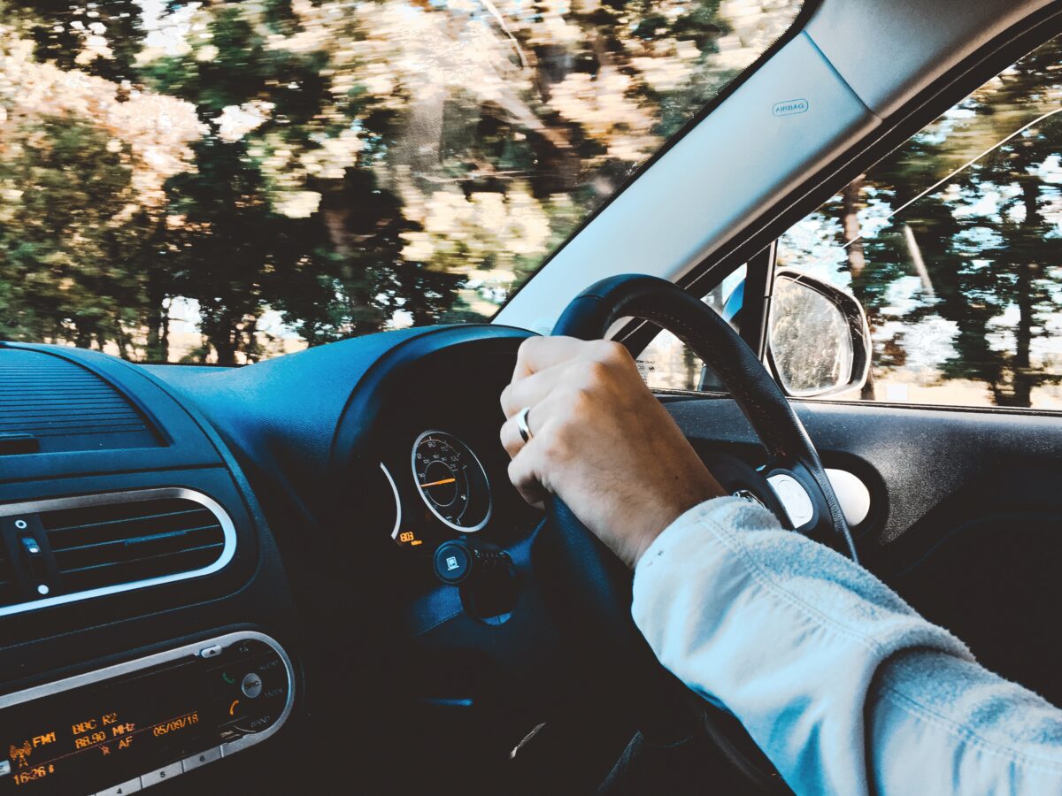 A driver's hand on the steering wheel of a car with the dashboard visible, driving through a sunlit, tree-lined road, possibly an Uber or Lyft.