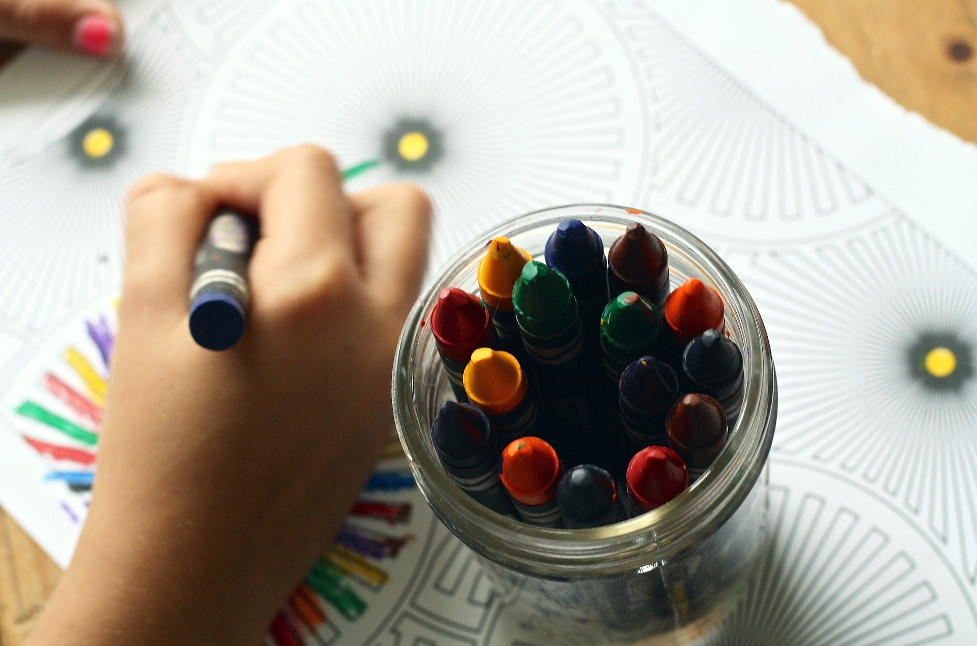 A child's hand holding a crayon over a coloring book with a glass jar full of colorful crayons on a wooden table at daycare, highlighting a creative activity setup focused on fostering artistic expression while ensuring