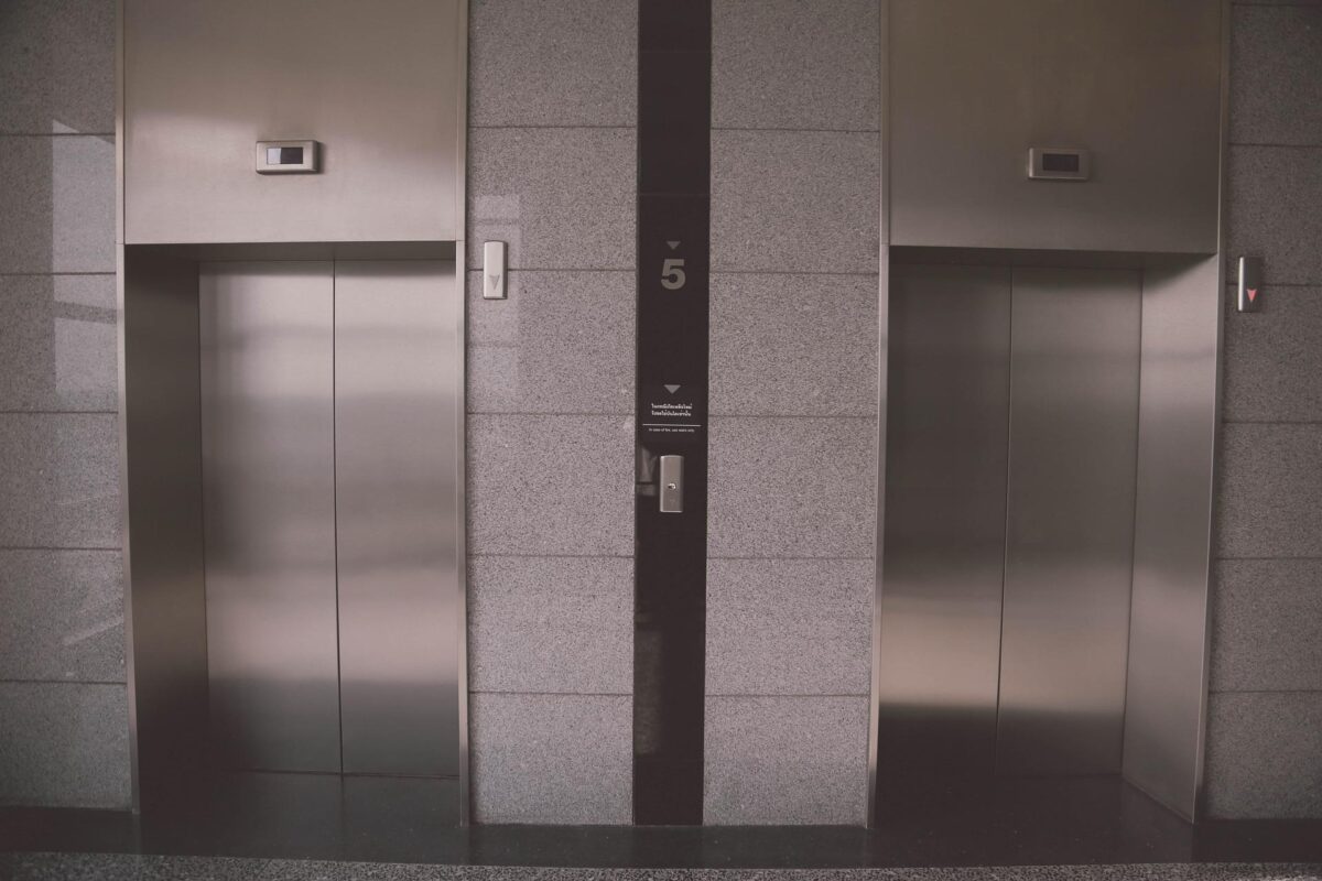 Three closed elevator doors on a gray tiled wall with a central call panel displaying the number 5, and emergency features indicated nearby to ensure safety against potential elevator accidents.