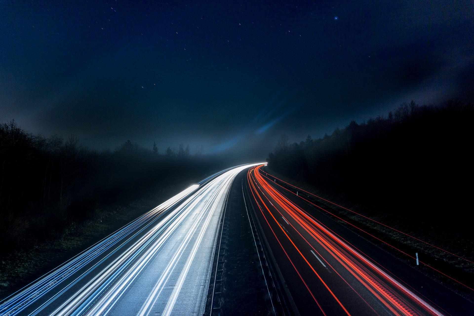 Long exposure shot of a highway at night, capturing streaks of white and red lights from speeding vehicles, surrounded by dark forest under a starry sky.