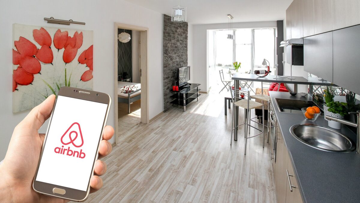 A hand holding a smartphone displaying the Airbnb accidents logo in a modern kitchen with a living room in the background, featuring a large floral wall decor and sleek furniture.