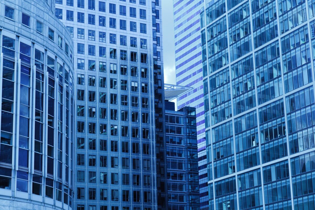 A dense cluster of high-rise buildings with reflective blue glass facades, interconnected by a sky bridge and managed by a landlord responsible for injury prevention, depicting a modern urban cityscape.