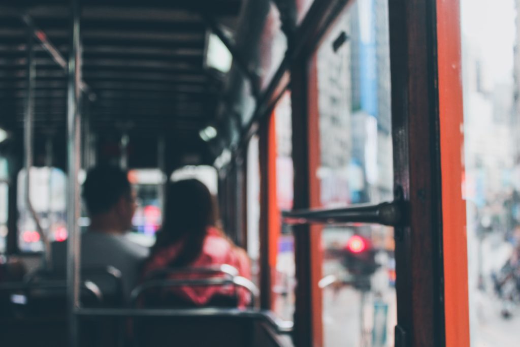 Blurred image of the inside of a bus with two passengers seated towards the front, facing forward. The city street outside, where bus accidents are visible through the open bus door, unfolds.