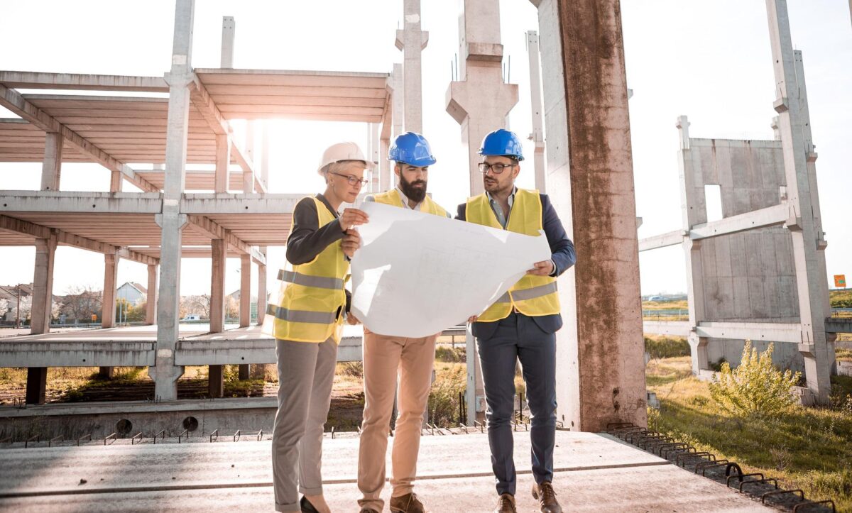 Three construction workers, two men and one woman, wearing hard hats and reflective vests, discuss a blueprint at a construction site with incomplete buildings in the background following a construction accident.