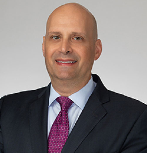 Professional portrait of Barry B. Ansbacher, a bald man in a dark suit, light blue shirt, and pink patterned tie, smiling at the camera. He has a confident and friendly expression