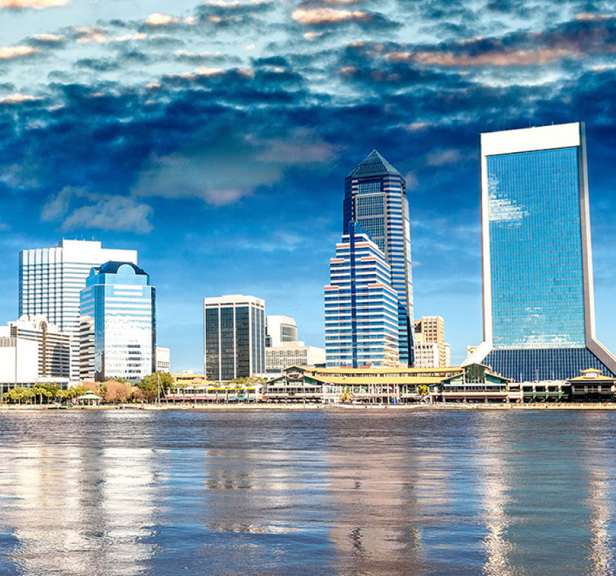 Skyline of a city with reflective high-rise buildings beside a body of water. The sky has dramatic clouds, and the water below mirrors the structures, enhancing the scene's vibrancy.