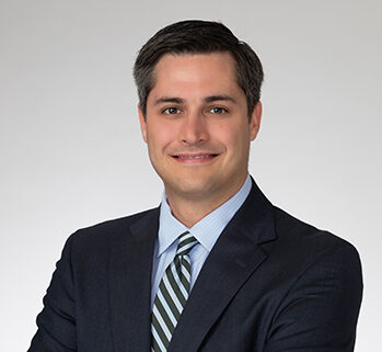 Portrait of a smiling professional man, Zachary R. Roth, in a blue suit and striped green tie against a solid, light gray background.