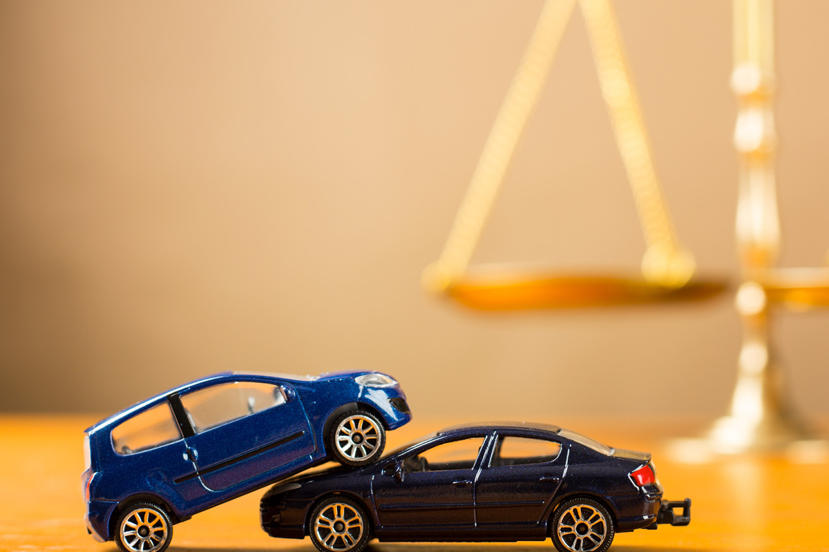 Two toy cars, one blue and one black, simulate a minor car accident in front of a blurred background featuring a scale of justice.