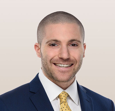 A professional headshot of Michael Feinberg, smiling with short hair, wearing a navy suit, white shirt, and yellow polka dot tie, against a light beige background.