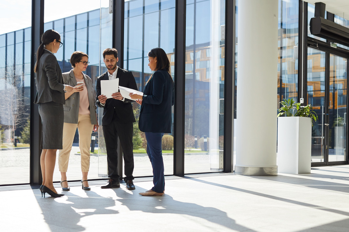 Four business law attorneys, two men and two women, engage in a discussion while holding documents in a modern office lobby with glass walls and a bright, sunny background.