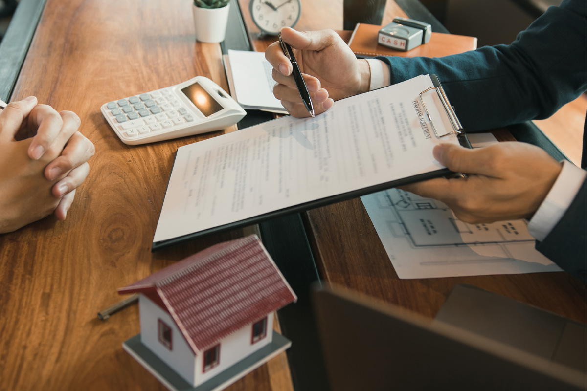 A professional meeting where two individuals, including a Commercial Real Estate Contract Attorney, discuss a document, with a calculator, small house model, and laptop visible on the wooden table.