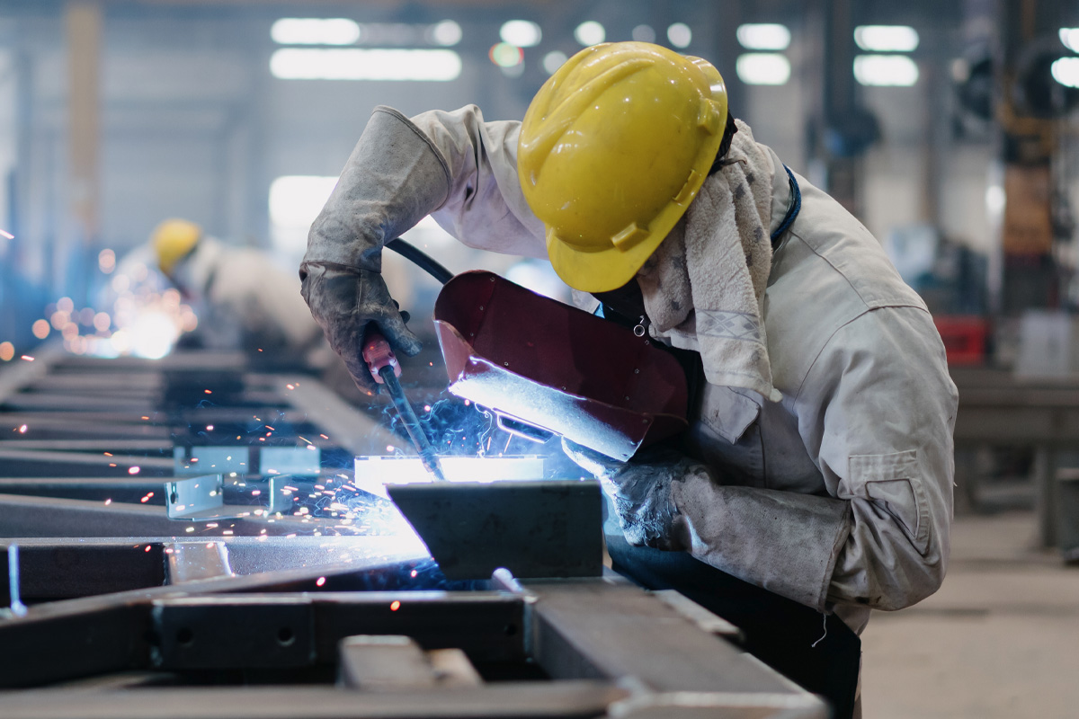A worker in a yellow hard hat and protective gear uses welding equipment on metal structures in an industrial setting. Sparks fly from the point of contact, and other workers can be seen in the background.