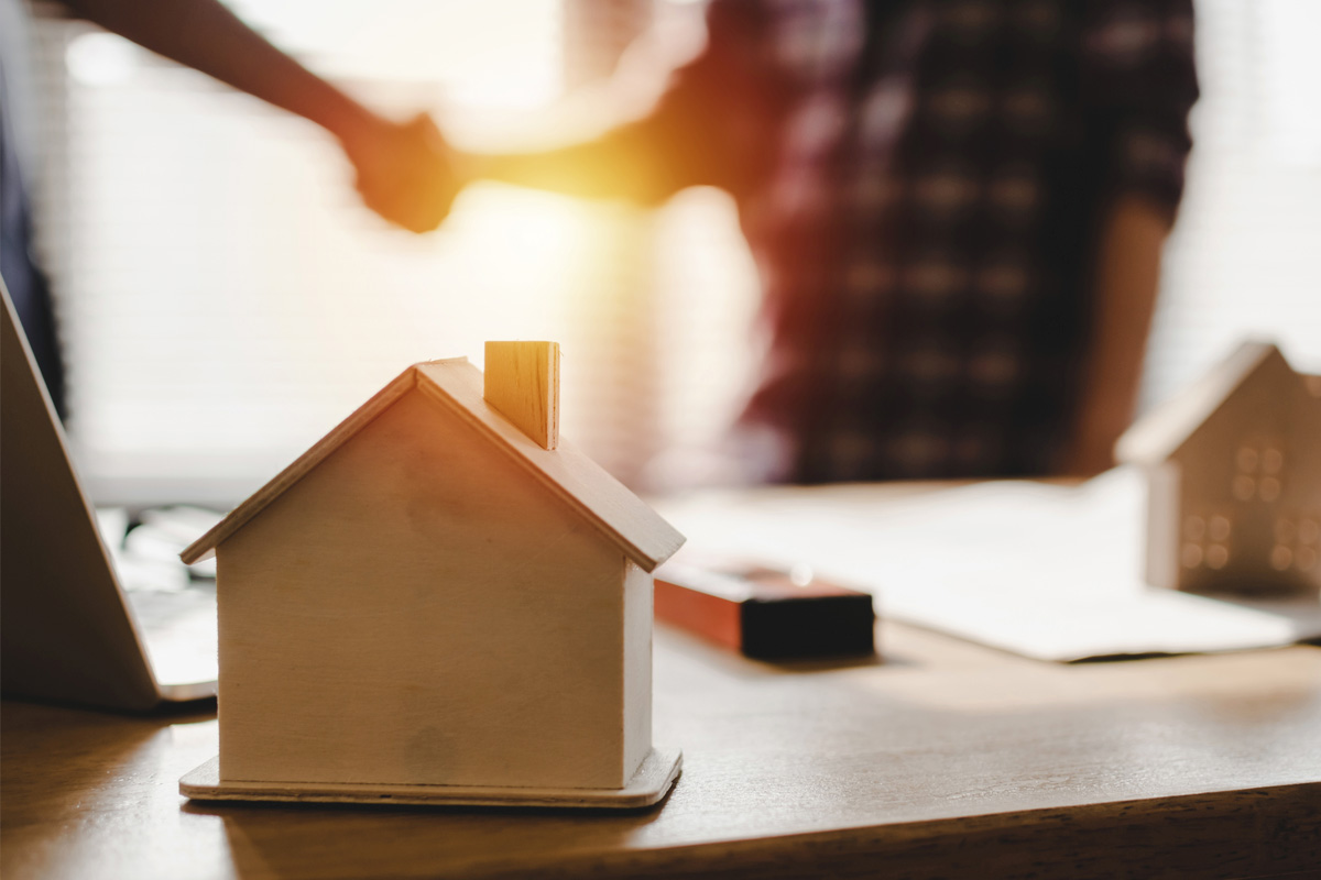A wooden model house on a desk with a warm, sunlit background where two construction lien attorneys are shaking hands, symbolizing a real estate transaction or agreement.