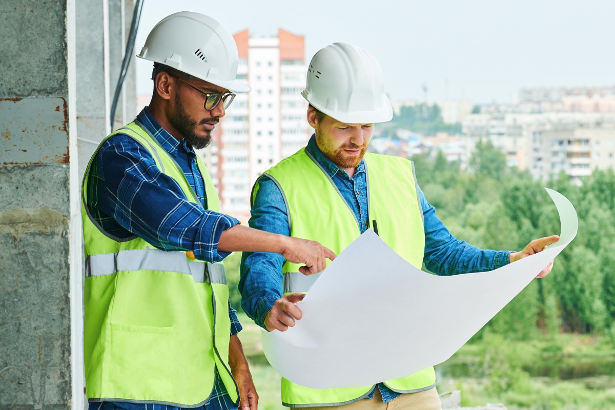 Two construction workers wearing white hard hats and yellow safety vests review architectural plans at a construction site. They stand near a partially built structure with a cityscape visible in the background.