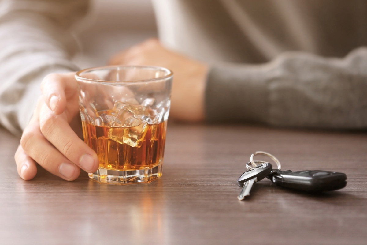 A person's hand rests near a glass of whiskey with ice on a table, with car keys placed close by, suggesting a narrative about drunk driving responsibility.
