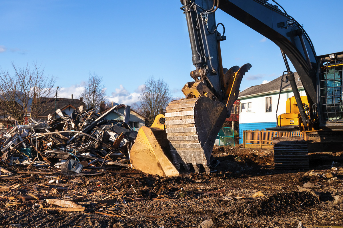 A large excavator clears debris at a construction site, with a pile of rubble and wood on the ground. A house can be seen in the background, under a clear blue sky.
