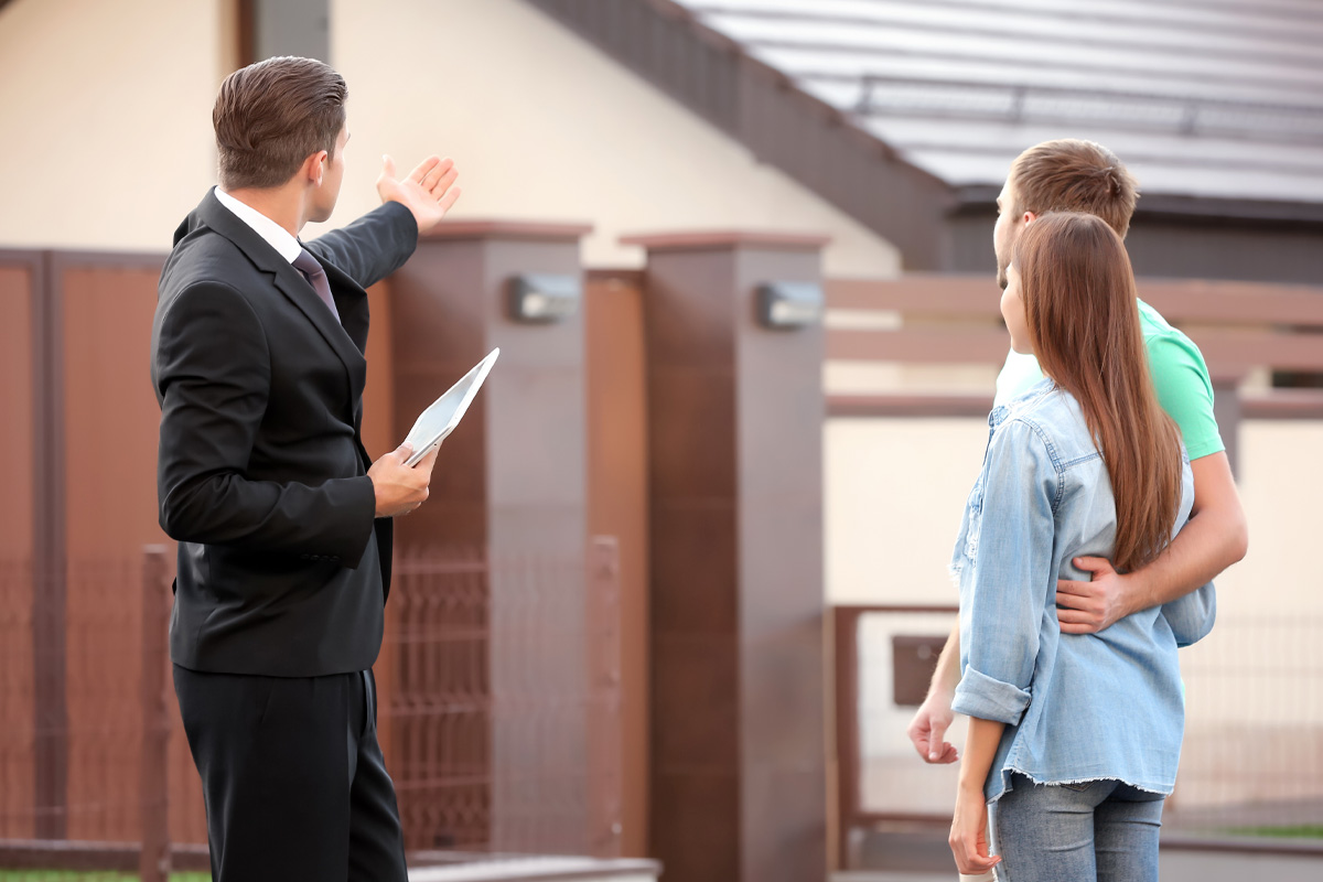 A real estate agent holding documents gestures towards a new house, as he shows it to a young couple standing nearby, viewing it together, potentially requiring the services of Failure to Disclose Attorneys.