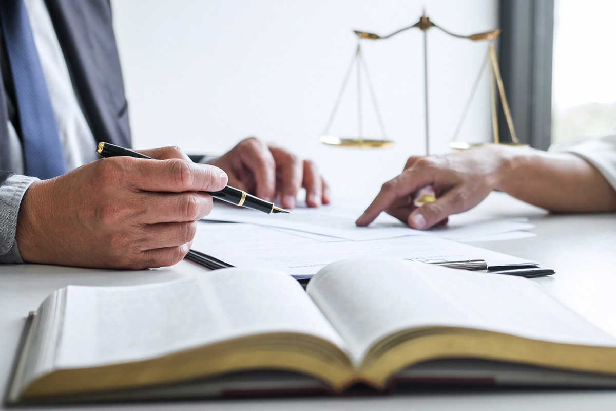 Two people are in a meeting, one pointing at papers and the other holding a pen. A large open book is in the foreground, and scales of justice are visible in the background, suggesting a legal or professional setting.