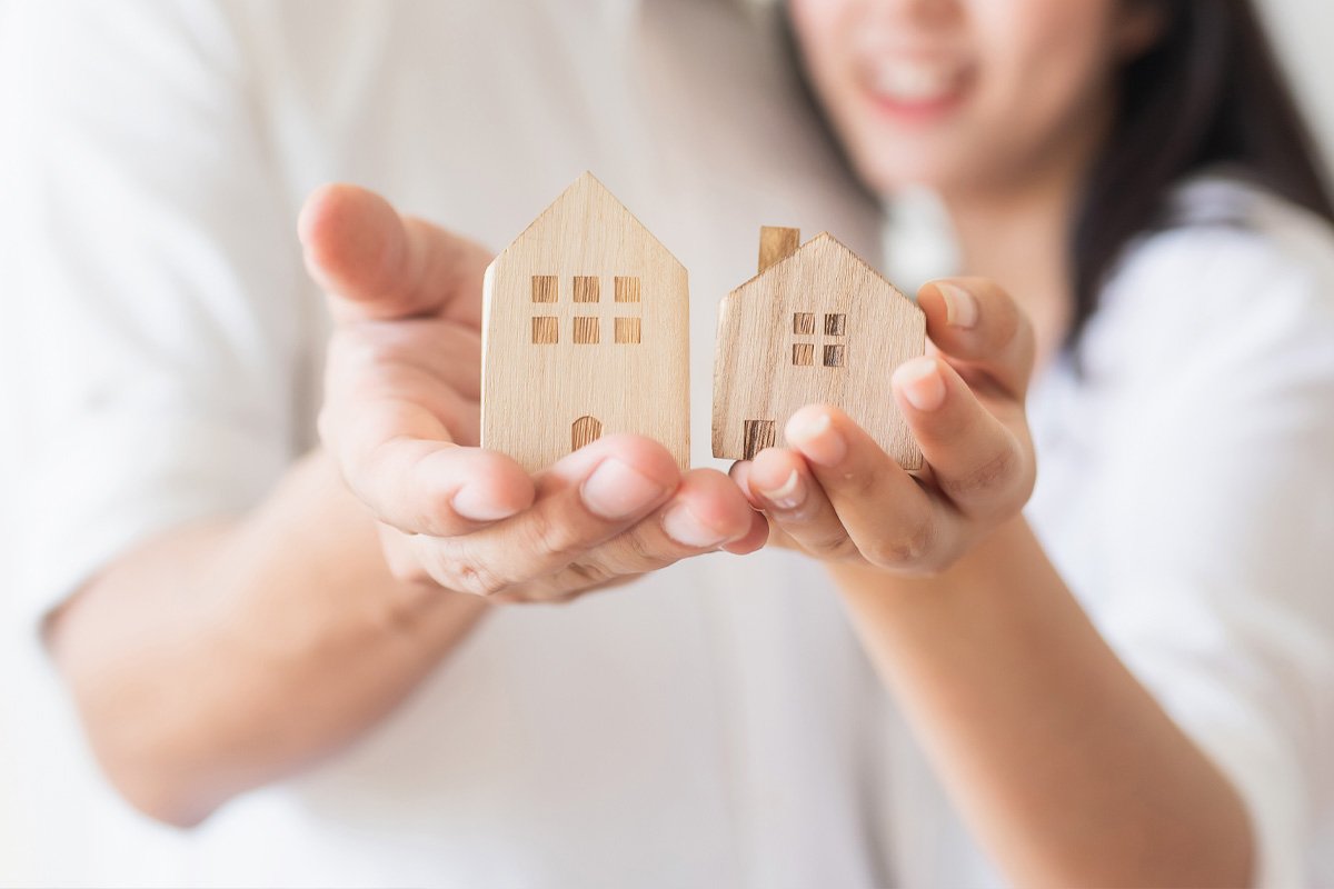 A smiling woman and a man holding two small wooden house models in their outstretched hands, symbolizing community home ownership or real estate investment.