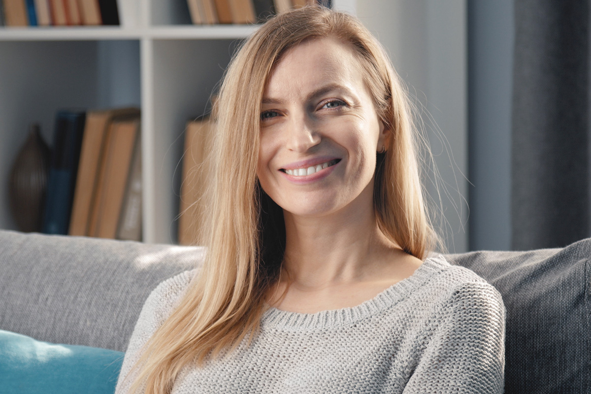 A smiling woman with long blonde hair sitting on a gray sofa in a cozy living room, with a bookshelf in the background. She wears a light sweater and looks relaxed and happy. Additional details