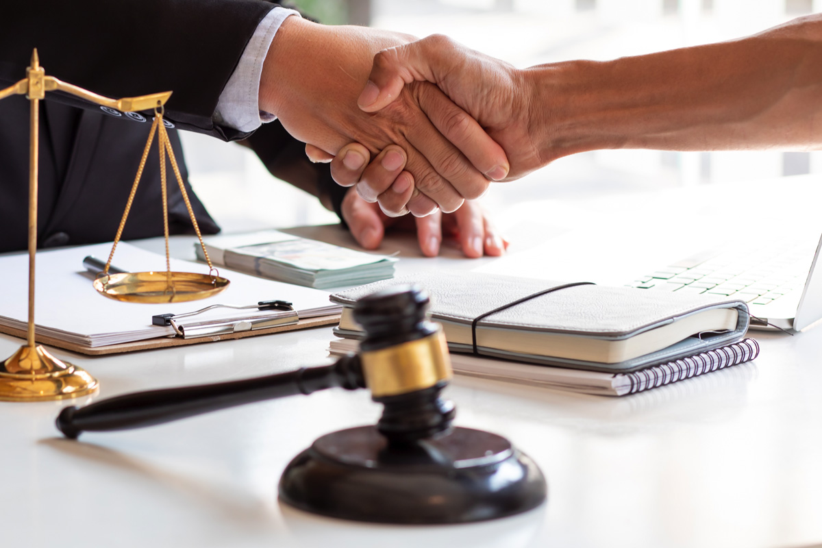 Two people shaking hands over a desk with a judge's gavel, scales of justice, documents, and a laptop, symbolizing a legal agreement or settlement.