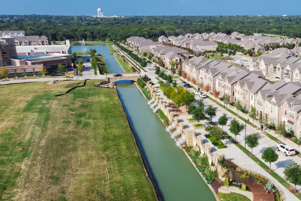 Aerial view of a suburban neighborhood featuring rows of townhouses alongside a canal, with a bridge crossing over and broad green lawns, under a clear sky, ideal for commercial real estate negotiation.