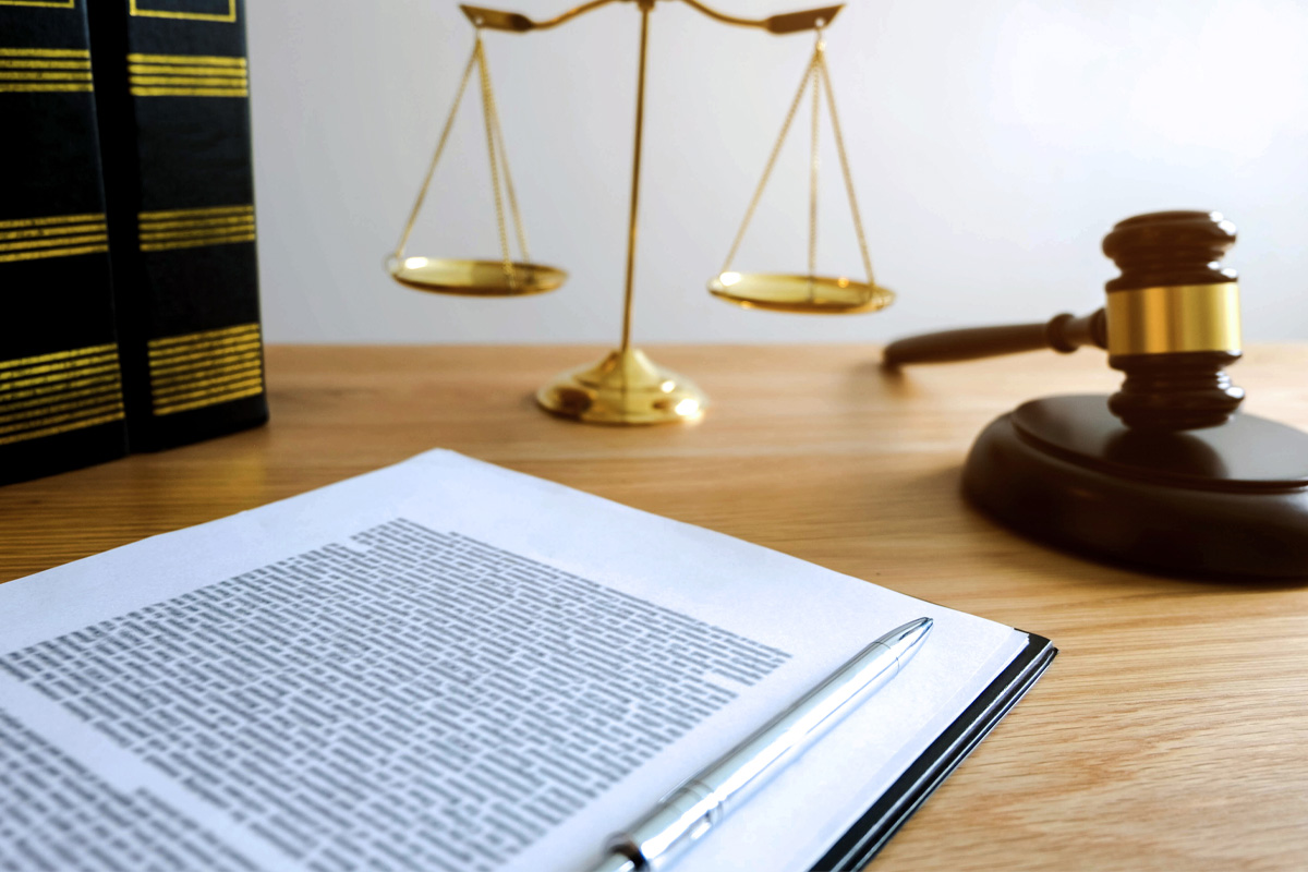 A wooden desk features a real estate contract, a pen, a gavel, and a set of scales, evoking a professional legal setting.