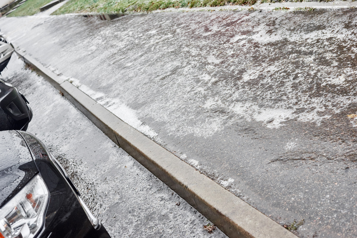 A snowy and icy residential street with visible tire tracks and an accident scene, showing accumulated snow on the curb and partly cleared asphalt, with a car's front corner visible on the left.