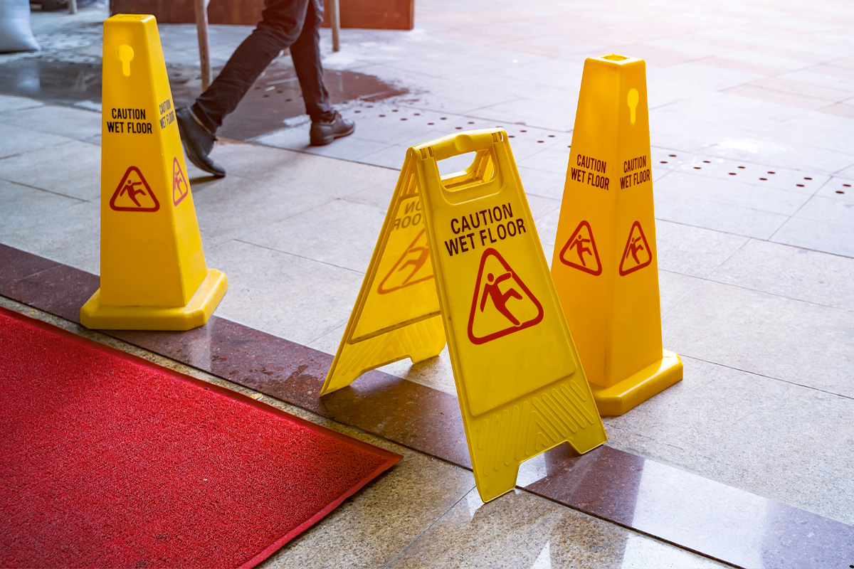 A person walks past several yellow "Caution: Wet Floor" signs placed on a marble floor, adjacent to a red carpet mat, as sunlight pours in from the right. It's a scene that would catch the eye of any Slip & Fall Accident Attorneys.