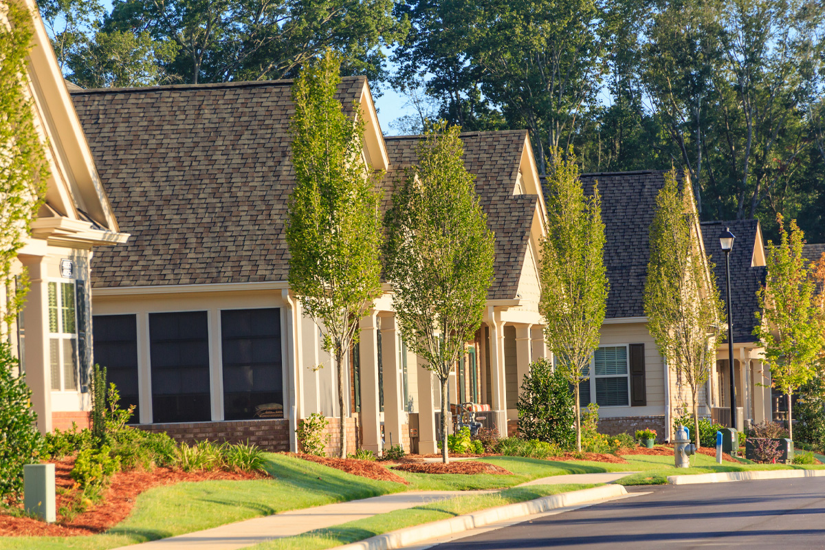 A row of modern suburban condominiums with gabled roofs, neatly maintained lawns, and young trees lining the sunlit street.