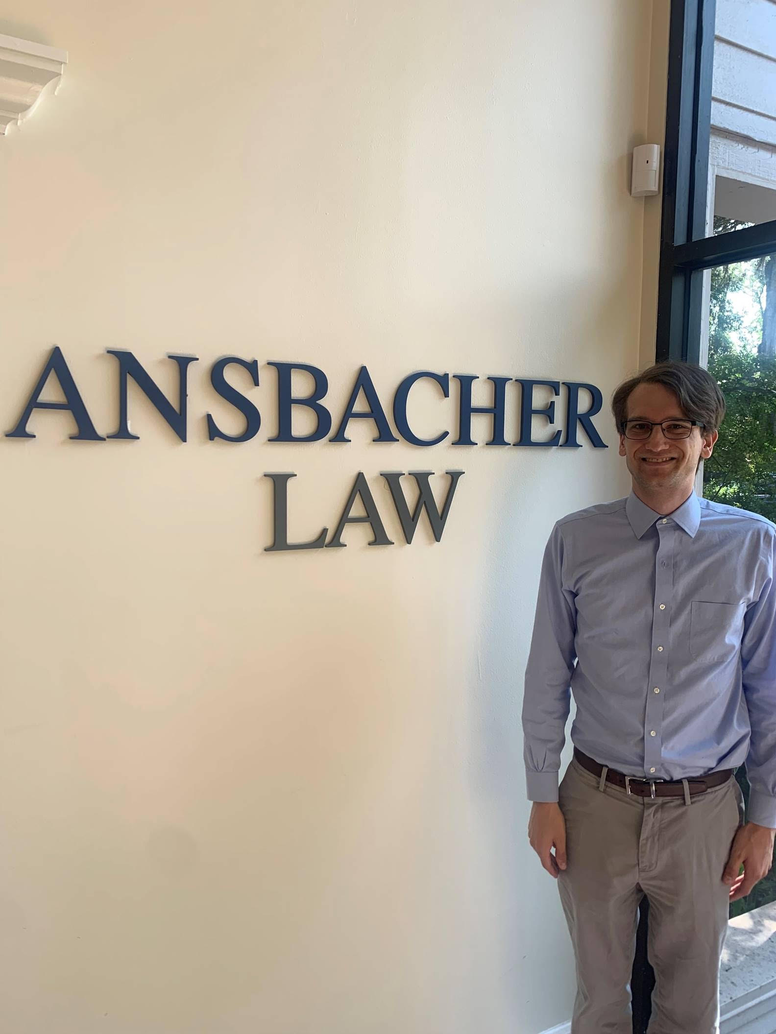 A person in a blue shirt and gray pants stands smiling next to a wall with the text "ANSBACHER LAW" in an office setting. Natural light is coming through a window on the right.