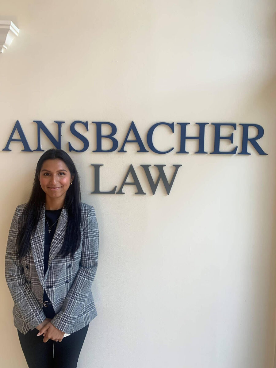 A woman in a gray blazer smiling in front of a wall with "Ansbacher Law" in raised dark blue letters.