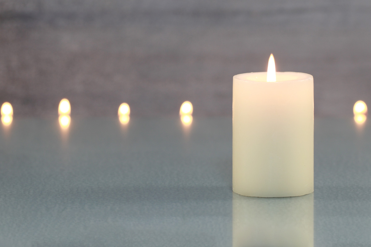 A single lit candle in the foreground with several smaller candles glowing in a blurred background, all placed on a reflective surface against a soft gray backdrop, symbolizing remembrance possibly used in wrongful death attorneys'