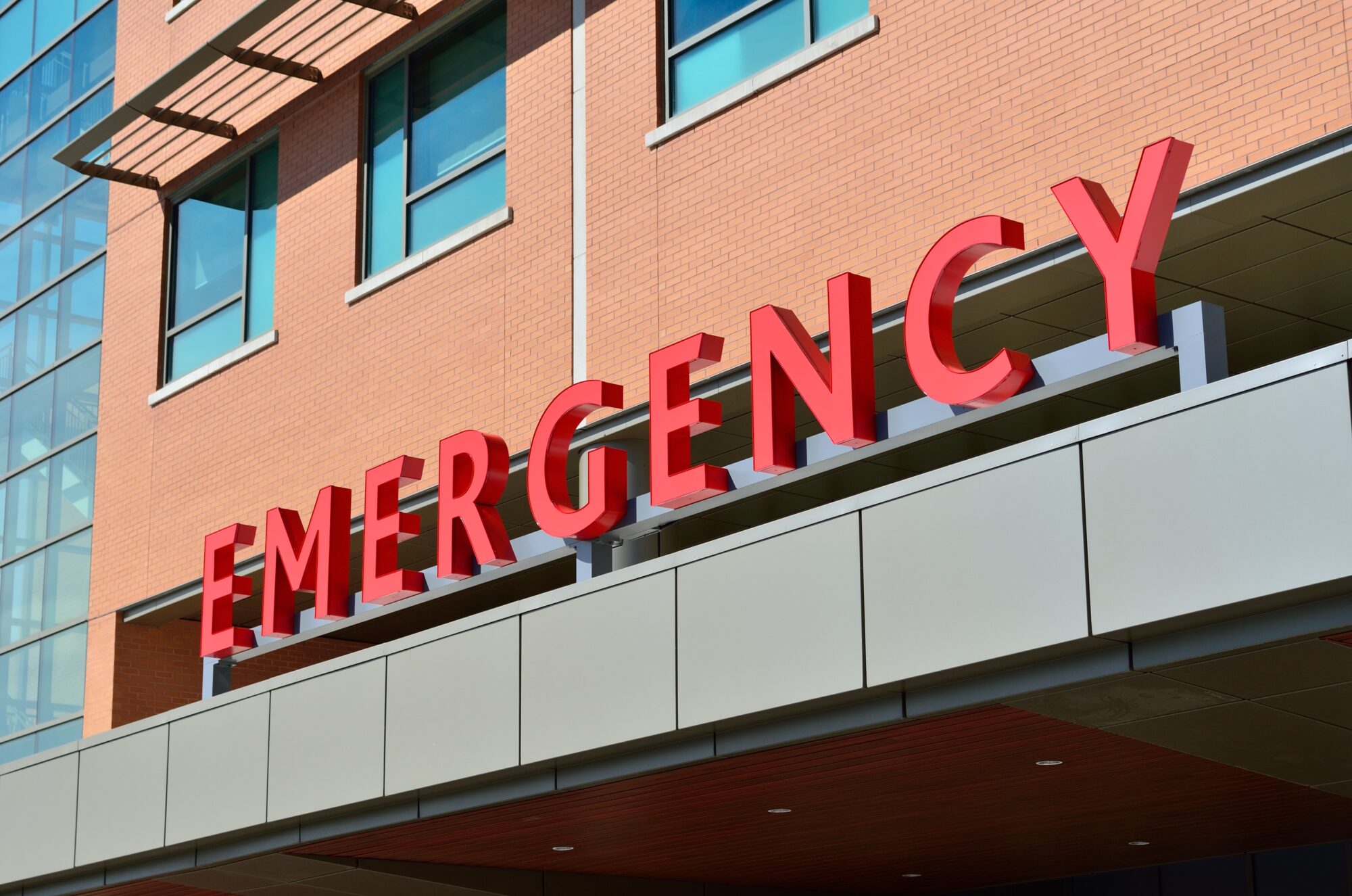 Red "emergency" sign on the facade of a modern hospital building specializing in defective product injury cases, with brick walls and large windows, under a clear sky.