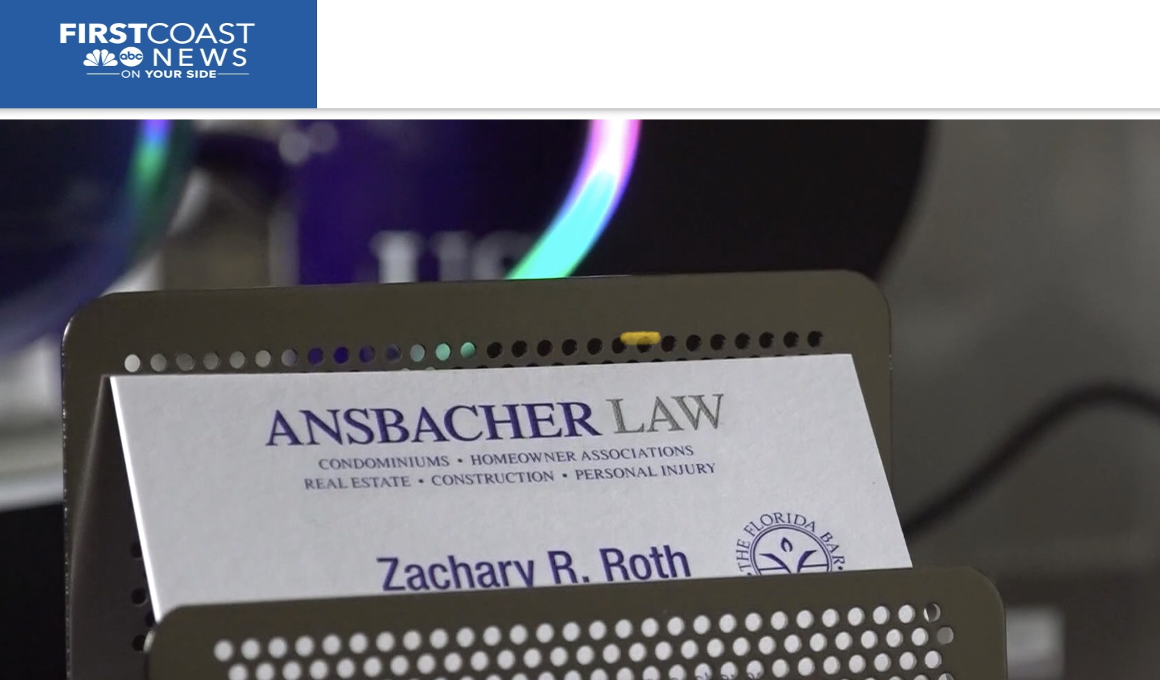 Business card of Zachary Roth from Ansbacher Law displayed in a card holder, with a blurred computer monitor showing the First Coast News logo in the background.